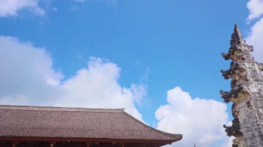 Geleneksel Bali tapınağının üzerinde tüylü beyaz bulutlar olan açık mavi bir gökyüzü. Tapınakta karmaşık taş oymalar ve sazdan bir çatı var. Bali mimarisi sergileniyor..