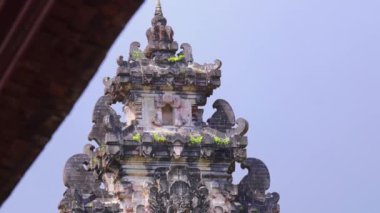 Geleneksel Bali tapınağı yapısına yakın çekim, karmaşık oymalar ve dekoratif öğeler sergileniyor. Tapınak yeşil yosunlarla süslenmiş ve açık mavi bir gökyüzüne kurulmuş..