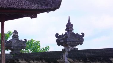 Duvarda ve çatı yapısında süslü taş oymalar bulunan geleneksel Bali mimarisinin yakın çekim görüntüsü. Sahne, arka planda yemyeşil yeşillikler ve bulutlu bir gökyüzünü içeriyor..