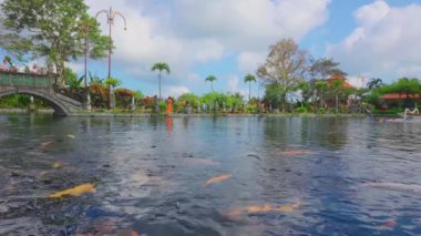 Renkli koi balıklarının yüzdüğü bir gölet içeren sakin bir manzara. Arka planda, bereketli yeşillik ve dekoratif bir köprünün yanı sıra manzaranın tadını çıkaran insanlar görülebilir. Gökyüzü parçalı bulutlu, sakin atmosfere ekleniyor..