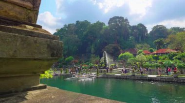 Ağaçlar ve geleneksel mimari ile çevrili yemyeşil bir bahçesi olan sakin bir manzara. İnsanlar su kenarında yürüyor, manzaranın tadını çıkarıyorlar..