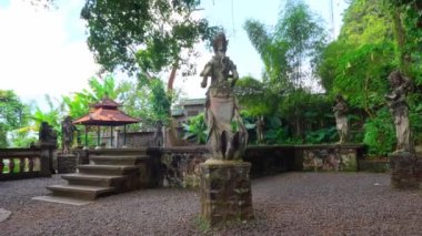 Serin bir bahçedeki taş heykellerin olduğu sakin bir açık hava sahnesi. Heykeller karmaşık bir şekilde tasarlanmış ve tropikal bitkilerle çevrili. Geleneksel bir çardak arka planda görülebilir ve sakin atmosfere katkı sağlar..