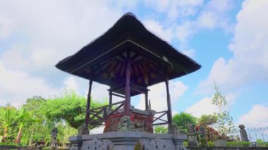 Yeşillik ve mavi gökyüzü ile çevrili sazdan çatılı geleneksel bir Bali pavyonu. Yapı karmaşık taş oymaları içeriyor ve sakin bir bahçe ortamında geçiyor..