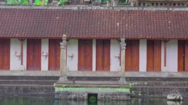 Huzurlu bir göletin yanına kurulmuş ahşap kapıları olan geleneksel bir Bali binasının huzurlu manzarası. Mimaride karmaşık taş oymalar ve yemyeşil çimlerle çevrili kırmızı kiremitli bir çatı yer alıyor..