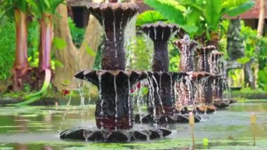 Bereketli yeşilliklerle çevrili çok katmanlı bir çeşmenin yer aldığı huzurlu bir bahçe sahnesi. Su, fıskiyeden yavaşça sakin bir gölete dökülür ve huzurlu bir atmosfer yaratır..