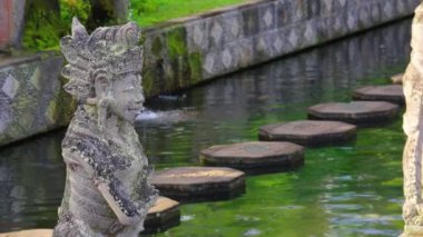 Sakin bir su yolunun yanında basamaklı taş bir heykelin olduğu sakin bir sahne. Heykel karmaşık bir şekilde oyulmuş, geleneksel kıyafetlerle betimlenmiş, yemyeşil ile çevrili..