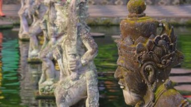 Arka planda su ve diğer heykellerle çevrili karmaşık detaylarla dolu taş bir heykelin yakından görüntüsü. Heykel yosun ve yıpranma içeriyor, yaşı gösteriyor. Ortam sakin ve kültürel görünüyor, muhtemelen bir tapınak ya da bahçede..