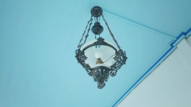 Mavi tavandan sarkan klasik bir avize. Karmaşık bir siyah metal işçiliği ve beyaz cam bir gölgesi var..