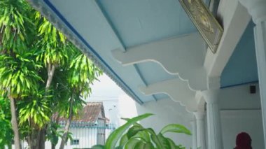 Mavi tavanlı ve beyaz mimari detayları olan huzurlu bir ev manzarası. Yeşil yemyeşil yapraklar ön planda ve arka planda görülebilir, barışçıl bir atmosfer yaratıyor..