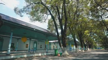 Bir tarafında geleneksel mimari olan, ağaçlarla kaplı bir patikanın huzurlu manzarası. Güneş ışığı yaprakların arasından süzülerek barışçıl bir atmosfer yaratır. Bölge temiz ve bakımlı, doğa ve yapının bir karışımını gösteriyor..