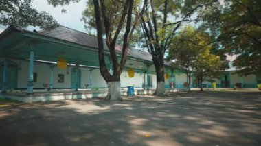 Görkemli yeşil ağaçlarla çevrili, mavi aksanlı geleneksel mimarinin yer aldığı sakin bir avlu. Alan, yaprakların arasından süzülen güneş ışığıyla aydınlanmış, barışçıl bir atmosfer yaratıyor..