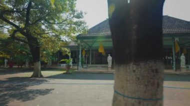 Geniş çatılı ve ağaçlarla çevrili geleneksel bir bina. Yapı sütunlara sahiptir ve asfalt yolu ile geniş bir alanda ayarlanır.