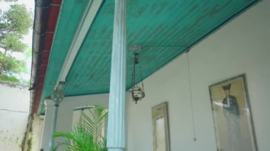 İçinde turkuaz bir tavan, asılı bir lamba ve duvarda iki çerçeveli portre bulunan eski bir odanın köşesi. Yeşil bir bitki iç kısımlara biraz doğa katar..