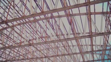 Büyük bir endüstriyel binanın çatı yapısını görmek, mavi gökyüzüne karşı metal kirişler ve kirişlerin karmaşık bir düzenini sergiliyor..