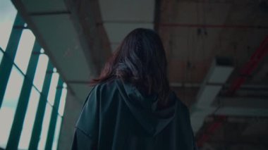 Geniş pencereli sanayi bölgesinde duran genç bir kadın, bol, siyah bir kıyafet giyiyor. Atmosfer dengesiz ve sanatsal, yumuşak ışıklandırma onun özelliklerini vurguluyor..