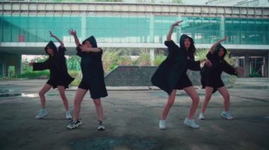 Dört dansçıdan oluşan bir grup dışarıda senkronize bir gösteri yapıyor. Uyumlu siyah kıyafetler giyiyorlar ve modern mimari bir zemine karşı dinamik hareketler sergiliyorlar..