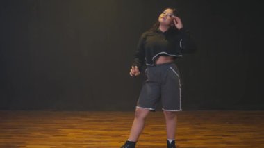 Kendine güvenen genç bir kadın dans stüdyosunda poz veriyor. Kırpılmış siyah bir kazak ve gri bir şort giyiyor. Arka plan koyu renkli, duruşunu ve tarzını vurguluyor..