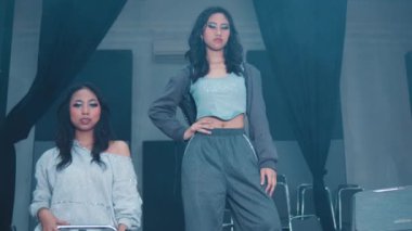 Bir grup altı genç kadın şık bir iç mekan ortamında poz veriyorlar. Moda kıyafetleri giyiyorlar, gündelik ve spor tarzlarının karışımını sergiliyorlar. Arka planda sandalyeler ve modern bir estetik, dramatik ışıklandırma sahneyi güçlendiriyor.