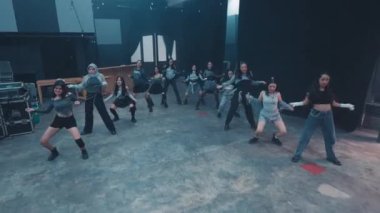Bir grup genç dansçı loş bir stüdyoda koreografi gösterisi yapıyorlar. Gri ve siyah tonlarında şık, günlük kıyafetler giyiyorlar, dans stillerinin karışımını sergiliyorlar.. 
