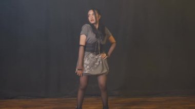 Genç bir kadın, pullu kısa gri bir elbise, korse kemeri ve siyah ayak bileği çizmeleriyle kendine güvenen bir şekilde poz veriyor. Arka plan karanlık, pozunu ve modasını vurguluyor..