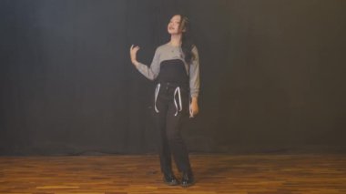 Genç bir kız, gri bluzu ve siyah pantolonu olan şık bir kıyafetle güven içinde poz veriyor. Arka plan karanlık, duruş ve ifadesini vurguluyor, bir dans ya da performans ayarını öneriyor..