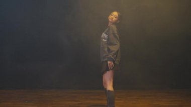 Genç bir kadın, üzerinde 'The Hamptons USA' yazılı gri bir kapüşonlu, kısa etek ve diz boyu çizmelerle güvenli bir şekilde podyumda yürüyor. Arka plan dumanlı, dramatik bir atmosfer yaratıyor..