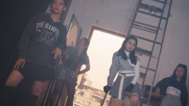 Loş ışıklı bir odada, arka planda bir merdiven ve açık bir pencereyle güvenle poz veren dört genç kadından oluşan bir grup. Şık kıyafetler giyiyorlar, gündelik ve modanın bir karışımını sergiliyorlar..