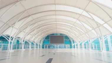 Geniş, modern kapalı alan, yüksek, kemerli tavan ve büyük boş zemin alanı. Tasarım beyaz yapısal kirişler ve uzak uçta büyük boş bir ekran içeriyor, minimalist bir estetik yaratıyor..
