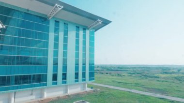 Pırıl pırıl tasarlanmış, yeşil tarlalar ve açık mavi gökyüzüyle çevrili modern cam bir bina. Bina, günışığını yansıtan ve çağdaş bir mimari stil sergileyen büyük pencerelere sahip..