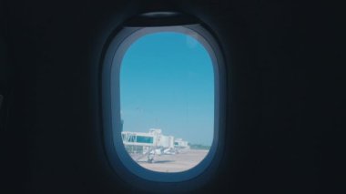 Açık mavi gökyüzünü ve havaalanı terminalini gösteren bir uçak penceresinden görüntüle.