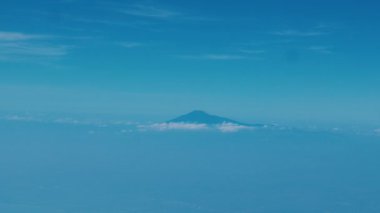 Açık mavi bir gökyüzünün altında bulutlardan oluşan bir denizden yükselen bir dağın uzak görüntüsü. Dağ bulutlar tarafından kısmen gizleniyor, sakin ve sakin bir atmosfer yaratıyor..