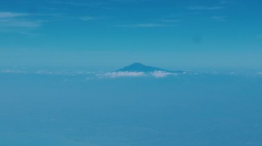 Bulutlardan mavi gökyüzüne doğru yükselen bir dağın havadan görünüşü. Aşağıdaki manzara sislerle kısmen gizlenmiş, sakin bir atmosfer yaratıyor..