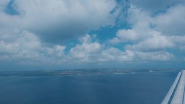 Mavi okyanuslu ve gökyüzünde dağınık bulutlu kıyı manzarası. Ufukta yeşil alanlar ve binalar görünüyor..