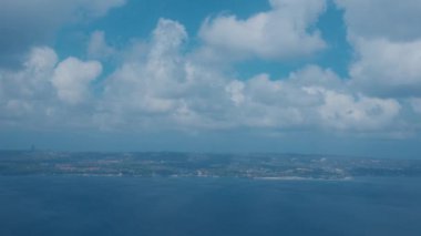 Mavi gökyüzünün altında, tüylü beyaz bulutlarla dolu bir kıyı şeridi manzarası. Okyanus gökyüzünü yansıtır, sakin bir atmosfer yaratır. Manzarada yeşil tepeler ve uzak binalar yer alıyor..