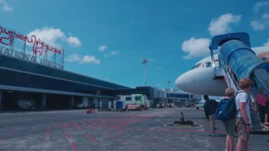 Yolcuların uçağa bindiği yoğun bir havaalanı sahnesi. Terminal binası üzerinde 