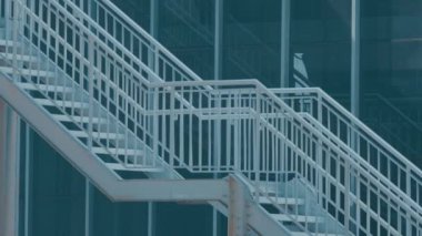 Camdan bir bina boyunca uzanan beyaz parmaklıklı modern bir merdiven. Tasarım parlak ve minimalistik, geometrik çizgiler ve şeffaflığı vurguluyor..