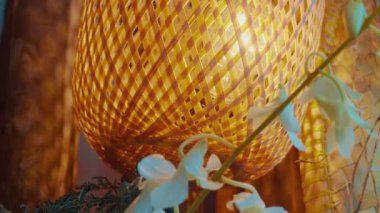 Narin beyaz çiçekler ve yeşil yapraklarla çevrili, içten parlayan sıcak bir bambu fenerinin yakın çekimi, sakin ve davetkar bir atmosfer yaratıyor..