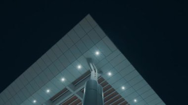 Geceleri modern bir binanın çatısının düşük açılı görüntüsü, geometrik bir tasarım ve aydınlatılmış ışıklar içeriyor. Yapıda parlak çizgiler ve çağdaş mimari stil sergileniyor.