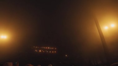 Karanlık bir atmosferi aydınlatan loş sokak lambaları olan sisli bir gece sahnesi. Arka planda binaların bulanık hatları var. Gizemli bir ortam yaratıyor..