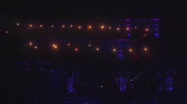 Loş ışık ve renkli spot ışıklarıyla dramatik bir sahne. Arkaplanda sanatçılar ve parlayan ışıkların siluetleri var, canlı bir atmosfer yaratıyorlar..