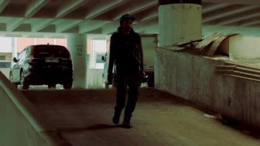 Loş bir otoparkta yürüyen siyah ceketli ve şapkalı bir adam. Çevre enkazla dolu ve arka planda park edilmiş bir araba var..