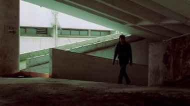 Yeraltı otoparkında yürüyen bir kişinin silüeti. Sahne sönük gölgeler ve beton duvarlarla aydınlatılıyor ve kasvetli bir atmosfer yaratıyor. Bu figür koyu renk giyinmiş ve bir şapka takmış. Gizemli bir hava katıyor..