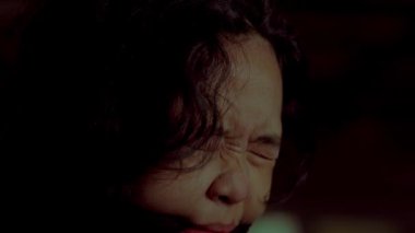 Gözleri kapalı, sakin bir yüz ifadesini gösteren bir kadının yakın çekimi. Saçları kıvırcık ve koyu renk ve ağzında siyah bir bez var. Işıklar loş, dramatik bir atmosfer yaratıyor..