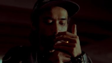 Uzun saçlı ve şapkalı bir adam sigara yakıyor. Sahne loş bir şekilde aydınlatılmış, kasvetli bir atmosfer yaratıyor. Odaklanmış ve derin düşüncelere dalmış görünüyor. Gölgeler onun özelliklerini vurguluyor..