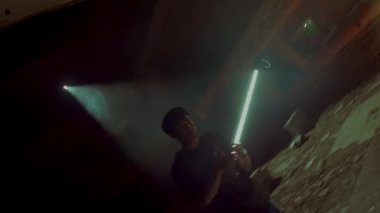 İki figürlü loş bir yeraltı alanı. Bir figür halatlarla bağlanırken, diğeri yakınlarda durup izliyor. Çevrenin cesur ve endüstriyel bir havası var. Işıklar ve gölgeler dramatik bir atmosfer yaratıyor..