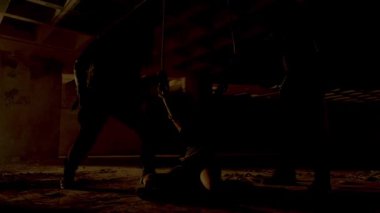 Terk edilmiş bir binada loş bir sahne, iki gölge figürün yer aldığı. Bir figür üzerinde bir çanta veya nesne asılı bir halat tutarken, diğer figür yakınlarda duruyor. Atmosfer gergin ve gizemli. 