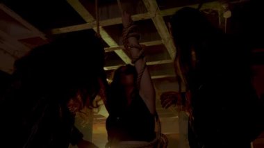 Terk edilmiş bir binada loş bir sahne, iki gölge figürün yer aldığı. Bir figür üzerinde bir çanta veya nesne asılı bir halat tutarken, diğer figür yakınlarda duruyor. Atmosfer gergin ve gizemli. 