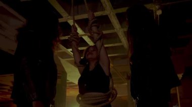 Terk edilmiş bir binada loş bir sahne, iki gölge figürün yer aldığı. Bir figür üzerinde bir çanta veya nesne asılı bir halat tutarken, diğer figür yakınlarda duruyor. Atmosfer gergin ve gizemli. 