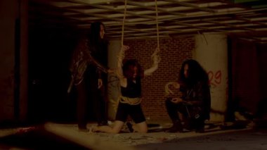 Terk edilmiş bir binada loş bir sahne, iki gölge figürün yer aldığı. Bir figür üzerinde bir çanta veya nesne asılı bir halat tutarken, diğer figür yakınlarda duruyor. Atmosfer gergin ve gizemli. 