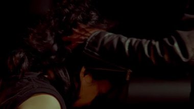 Uzun siyah saçlı birinin deri ceket giyen biri tarafından zapt edildiği dramatik bir sahne. Işıklandırma düşük, gergin bir atmosfer yaratıyor. İki birey arasındaki etkileşime odaklanıyoruz.